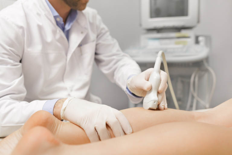 Arzt in Kittel und Handschuhen fährt mit Ultraschallsonde über den Unterschenkel eines Patienten.