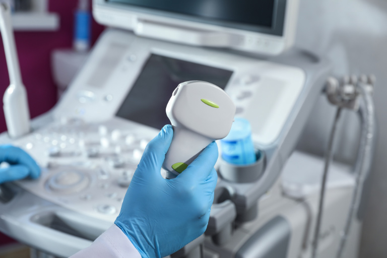 Person mit blauen Gummihandschuhen steht an Ultraschallgerät und hält Ultraschallsonde.